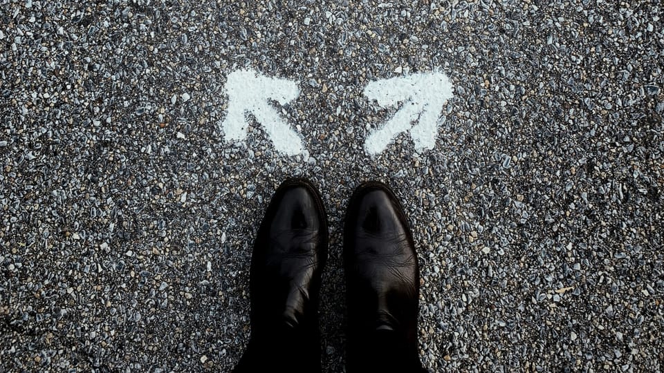A pair of dark leather shoes on coarse asphalt, facing two white arrows painted on the ground—one veering left, the other right—suggesting a decision or choice of direction.